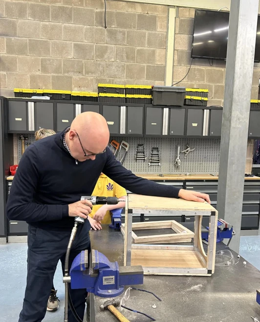 Man using a drill on a wooden project in a workshop at Glasgow Kelvin College, surrounded by tools and equipment. Man using a drill on a wooden project in a workshop at Glasgow Kelvin College, surrounded by tools and equipment.